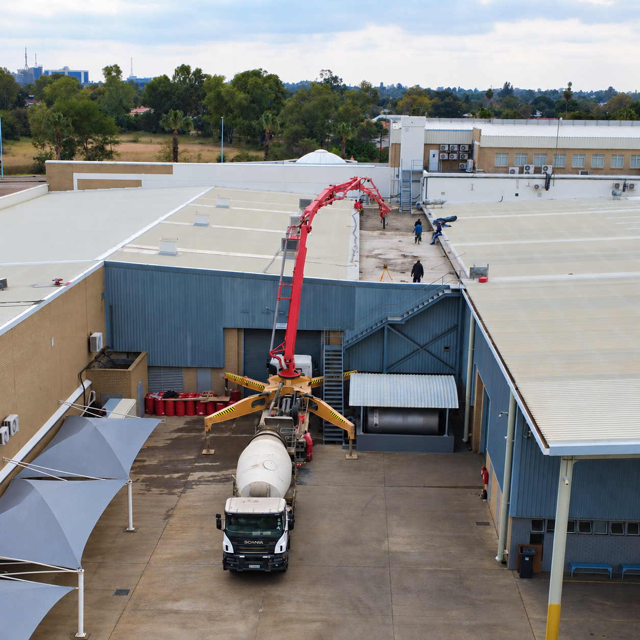 Overhead view of a Scania concrete mixer truck with a red boom pump extended to a roof at an industrial site